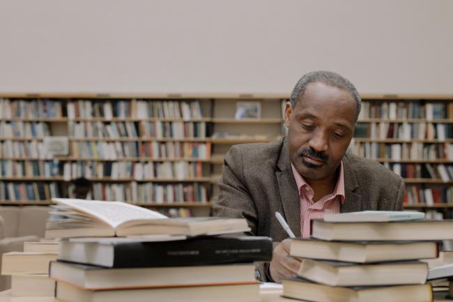 A professor focuses on their writing, surrounded by a pile of books on the table
