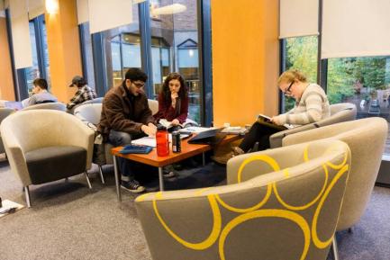 Students siting near the large windows of the skiybrdige in curved soft chairs around a table covered in water bottles, books, and papers. One student is reading a book on their lap.