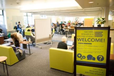Bright indoor area filled with green furniture, tables, chairs, whiteboards, and people in blurred focus. Large sign that reads "Hacherl Research & Writing Studio, WELCOME!"