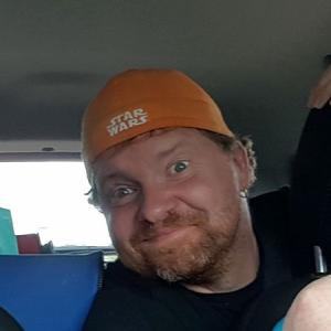 Ron is wearing an orange baseball cap backwards on his head and a dark t-shirt. He's leaning to the left, looking into the camera with a goofy grin on his face.