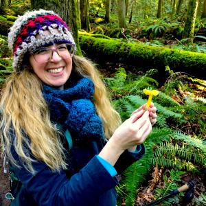 woman in hat and scarf in a forest holding a yellow mushroom