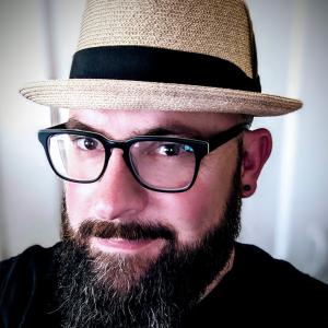 Head shot of Brian, a white male with a full brown beard gently smiling, wearing glasses and a tan fedora