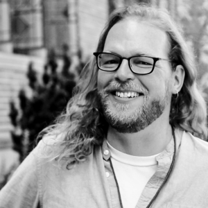 Black and white photo of Gabe--a white man with glasses, long hair, and a beard--smiling in front of the Wilson Library building..