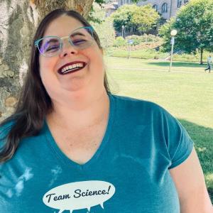 Jenny is standing in front of a tree, laughing. She's wearing a turquoise "Team Science" t-shirt, aqua glasses, and red lipstick. Old Main is in the background.
