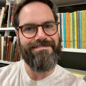 David is in front of colorful Yiddish periodicals. He wears glasses, has dark hair, light skin, and a salt-and-pepper beard