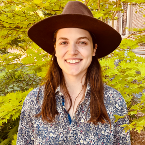 Portrait of Anika smiling and wearing a floral print shirt and wide brimmed brown hat in front of a vine maple tree.