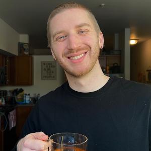 Brendan is wearing a navy crewneck sweater holding a glass mug with tea. He is smiling looking directly into the camera. He has a buzzed blonde head, and blue eyes. 