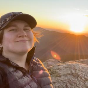 Emily on the summit of a mountain at sunset smiling and wearing a hat and a puffy coat.