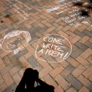 Brick sidewalk with chalk drawing of a face and speech bubble saying, "Come Write a poem!" and part of a written poem in chalk 