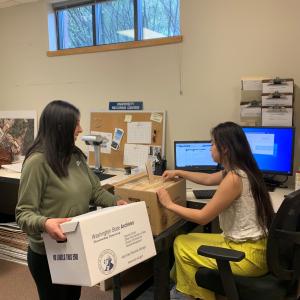 Two student employees working for University Archives & Records Management in an office in the Goltz-Murray Archives Building. One student holds an archival box while the other sorts through archival folders in another box. 