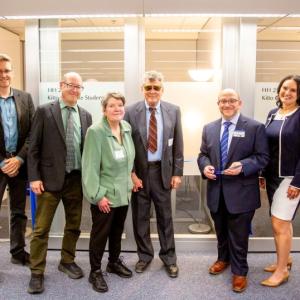 A group of people standing in front of the blue ribbon tied between the glass consultation rooms of the new Hub, moments before the ceremonial ribbon cutting. From left to right: Brad Johnson, David Patrick, Kathy Kitto, Dennis Waller, John Danneker, and Kim O’Neill.