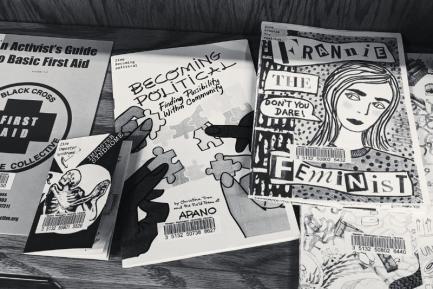 Black and white photo of several zines on a shelf with titles like: "Imposter Syndrome," "Becoming Political: Finding Possibility Within Community," and "Frannie the Feminist."